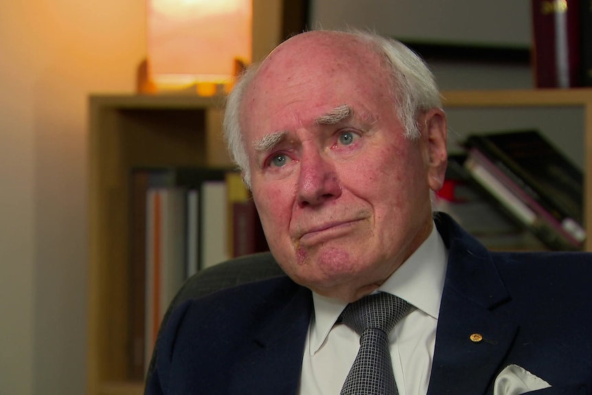 John Howard wearing a suit and tie sitting in front of a bookcase looking to the left