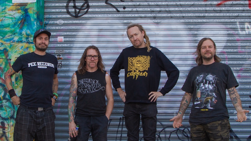 Four members of Australian punk band Frenzal Rhomb stand in front of a closed, graffitied garage door