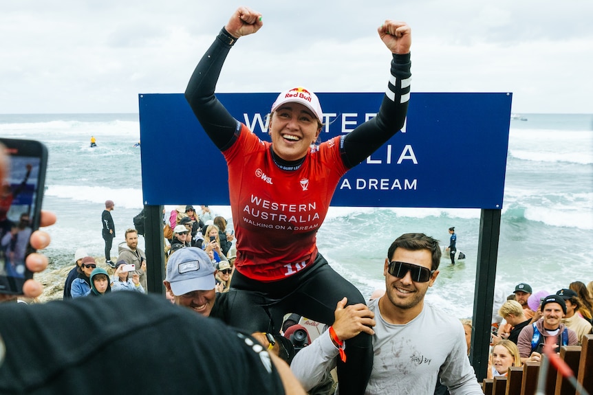 Carissa Moore lifts her arms in the air in celebration as sits on two men's shoulders