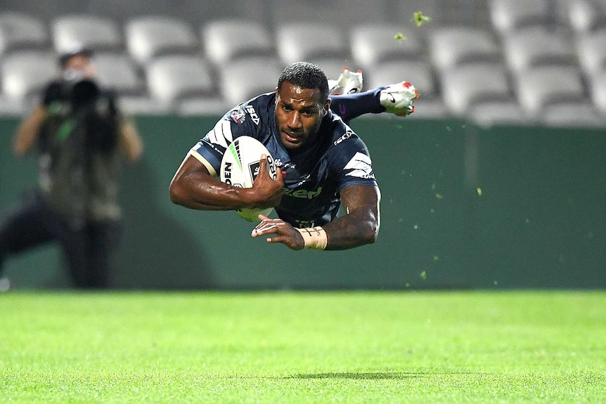 A Melbourne Storm NRL player divers in the air with the ball held in his left arm as he is about to score a try.