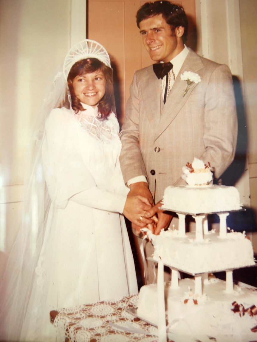 A couple, dressed in a white bridal dress and a fawn suit, are about to cut their three-tier wedding cake