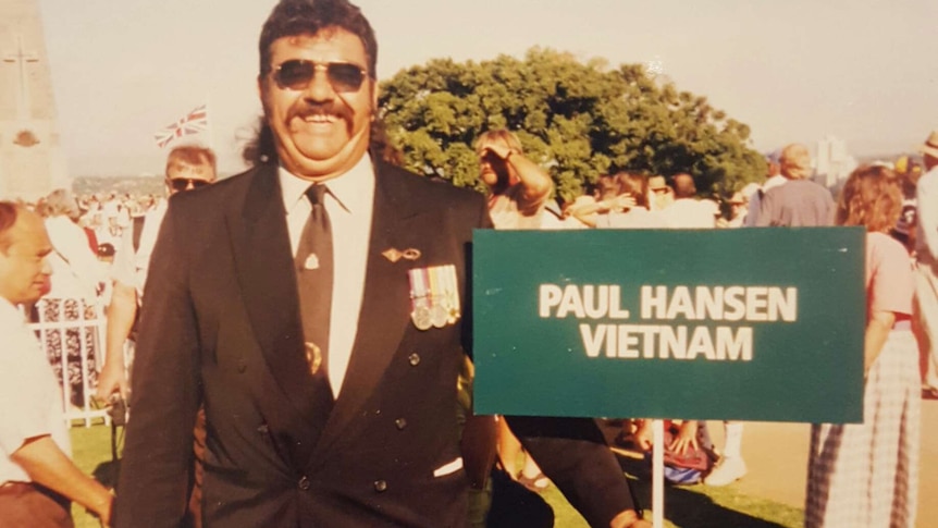 A man in a suit stands holding a green sign reading 'Paul Hansen, Vietnam'.