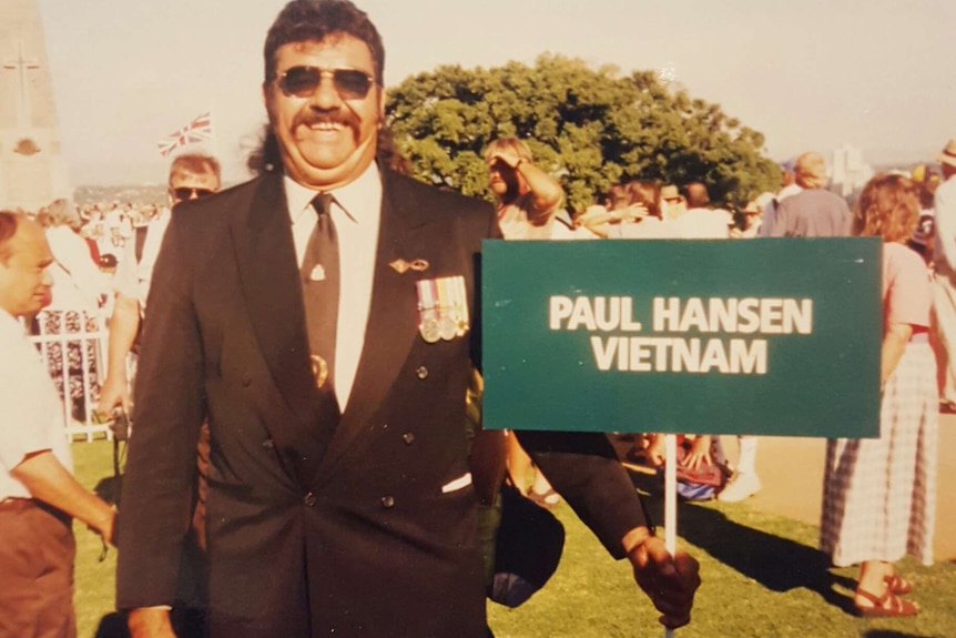 A man in a suit stands holding a green sign reading 'Paul Hansen, Vietnam'.