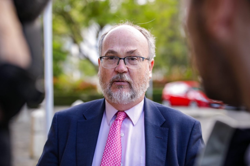 A middle-aged man with a grey beard and glasses, wearing a blue suit jacket with a pink tie faces the media.