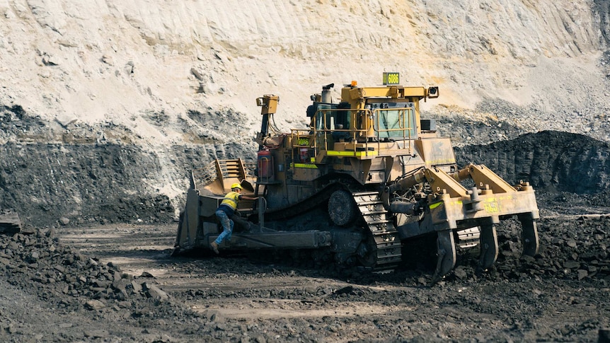 A man leans over the back of a machine that looks like it is used to break rocks. It sits in a large coal pit.