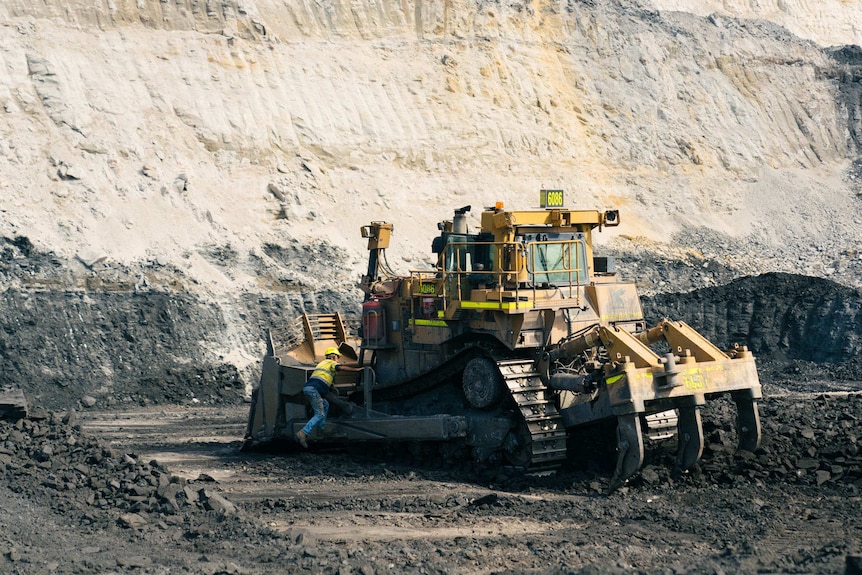 A man leans over the back of a machine that looks like it is used to break rocks. It sits in a large coal pit.