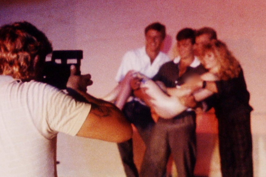 Three men hold a naked woman, while a male photographer takes their picture. The photo subjects are all smiling.