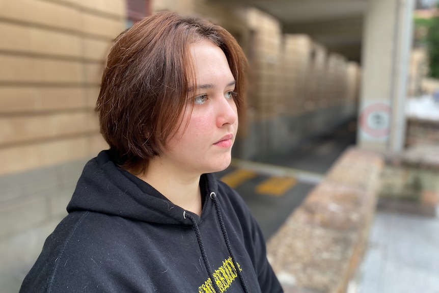 Rhiannon, wearing a black hoodie, sits in front of a brick building. 