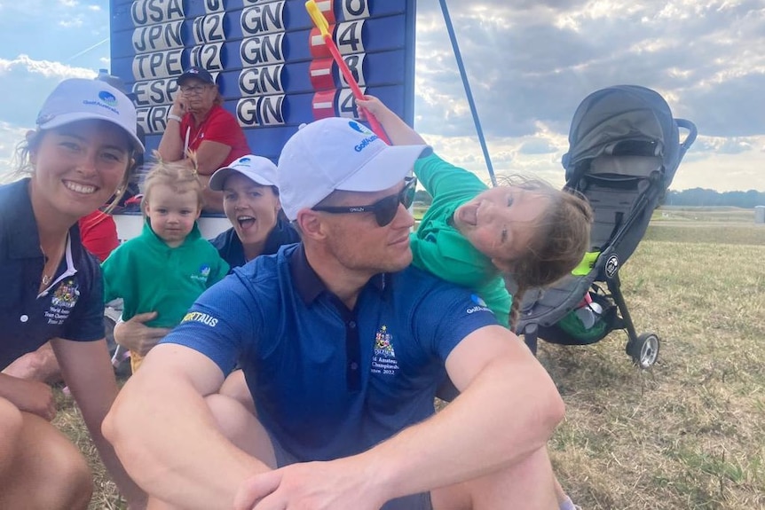 Kids climb on a man who's sitting on the ground in front of a golf scoreboard.