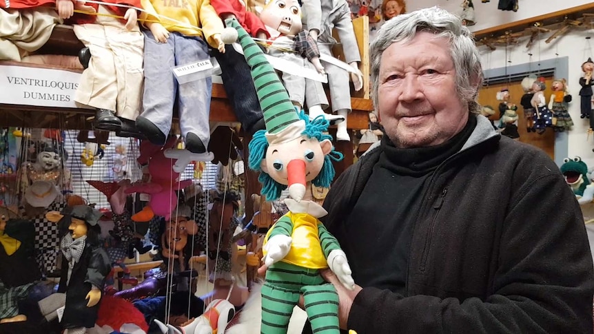 An elderly man with a Mr Squiggle puppet in front of a shelves filled with a large puppet collection.