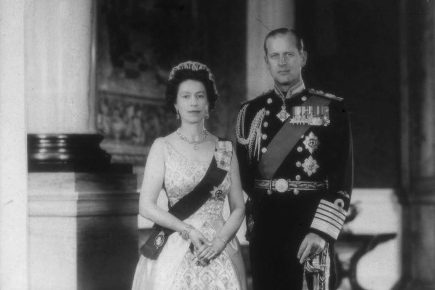 Woman wearing a crown standing next to older male with uniform covered in medals.