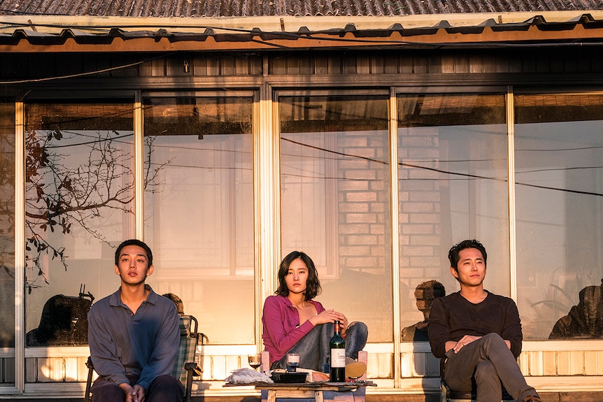 Colour still of Yoo Ah-in, Jeon Jong-seo and Steven Yeun sitting and watching sunset in 2018 film Burning.