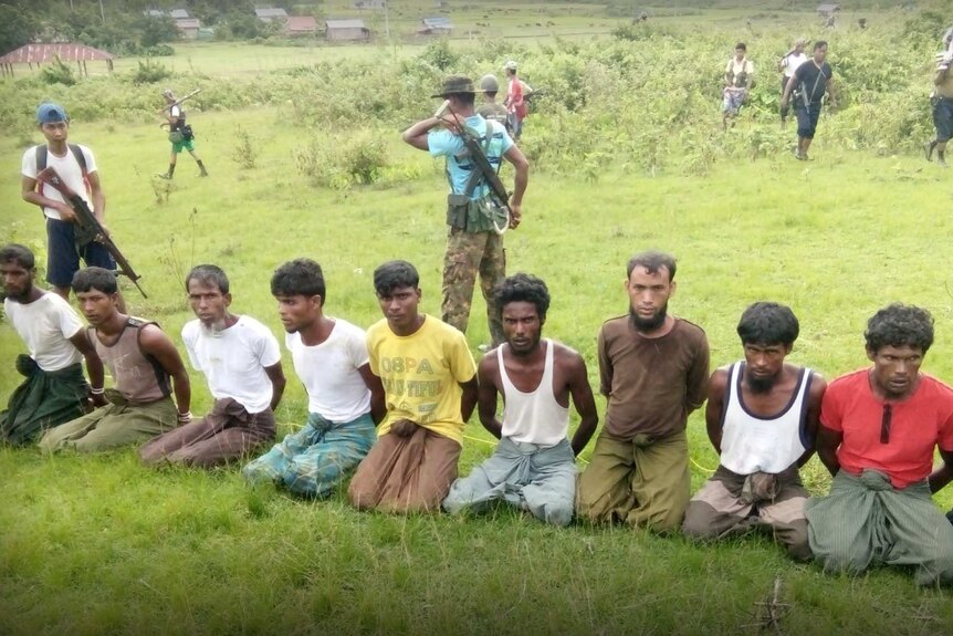 Wide shot of 10 men on their knees with their hands bound and armed men standing behind them.
