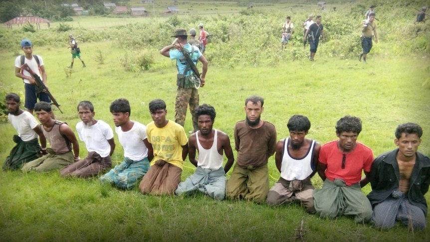 Wide shot of 10 men on their knees with their hands bound and armed men standing behind them.