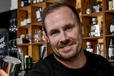 Scott Imlach holds up a pint of beer behind a bar smiling at the camera