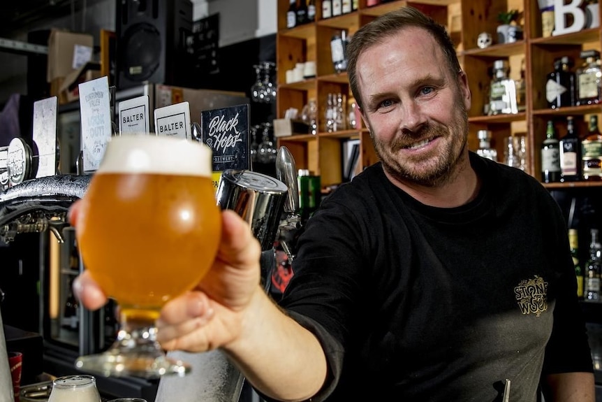 Scott Imlach holds up a pint of beer behind a bar smiling at the camera