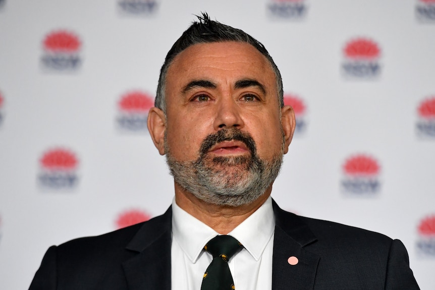 An olive-skinned, grey-bearded man wearing a dark suit and tie speaks in front of a government media conference backdrop 