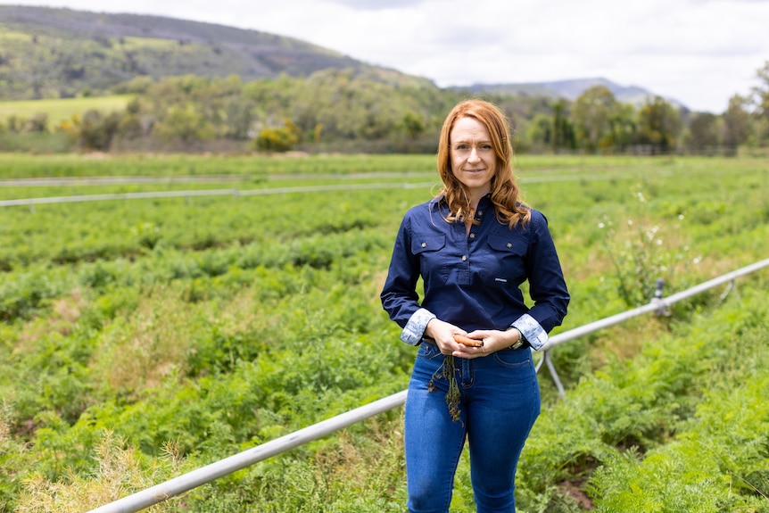 A woman stands in a field with irrigation pipes and mountains behind her.