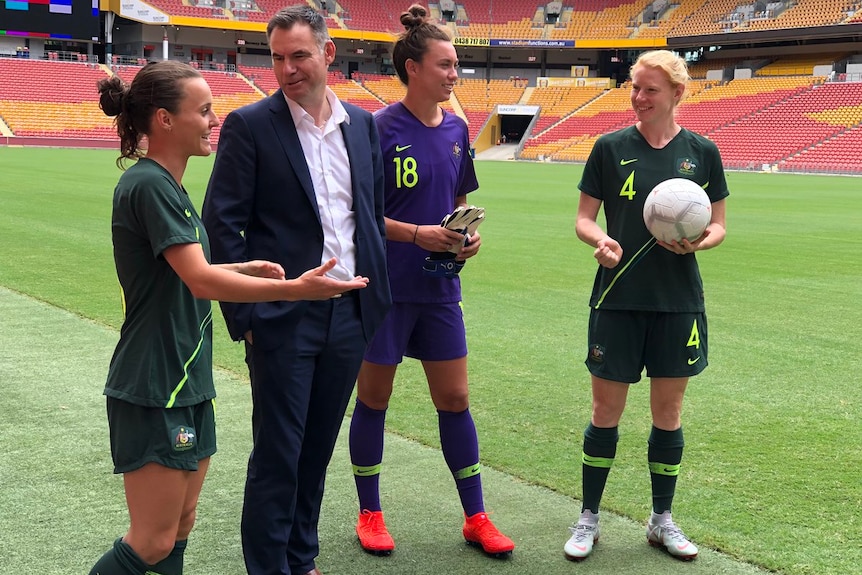 Ante Milicic stands next to the Lang Park playing surface speaking to three Matildas players.