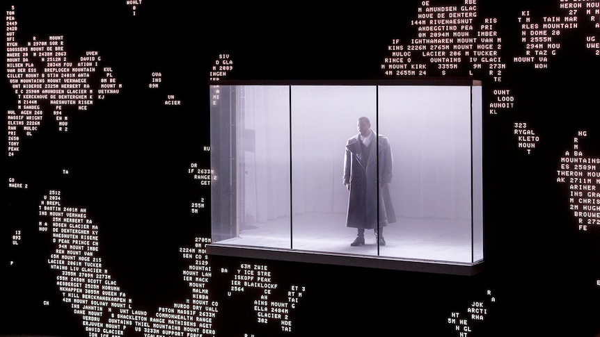 A man stands in a glass box on a stage, the backdrop is made up of data code.