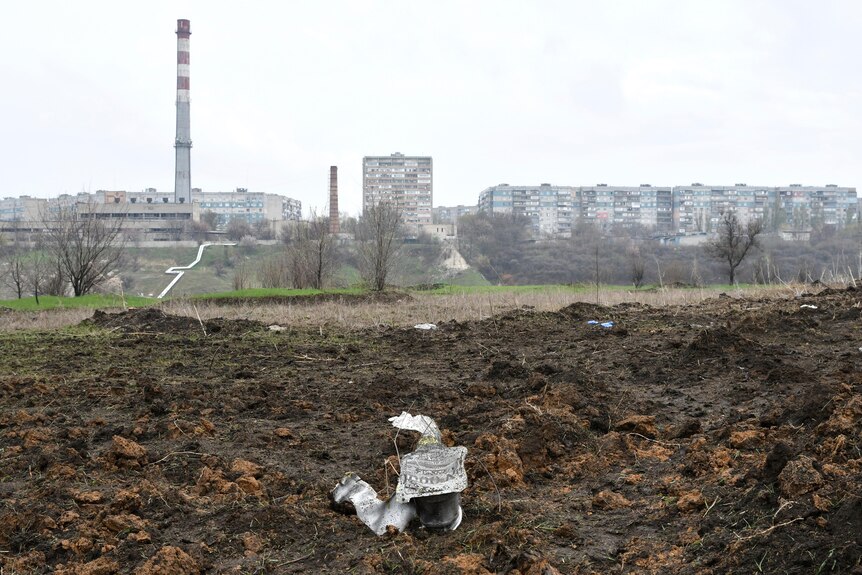 A part of a missile lies on the ground in front of an industrial landscape