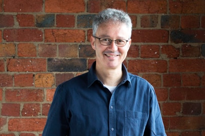 A man with blue shirt and orange pants stands in front of a red-brick wall, smiling slightly.