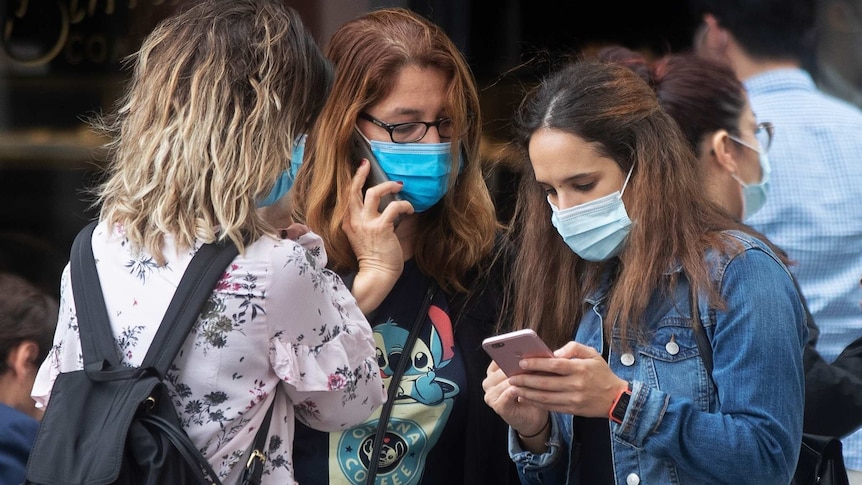 three young women wearing masks standing together and using their phones