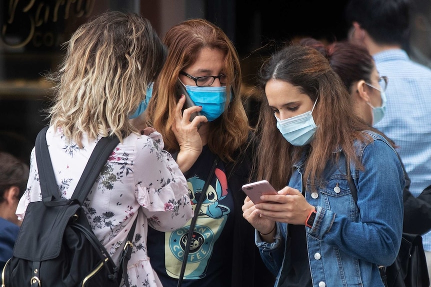 three young women wearing masks standing together and using their phones