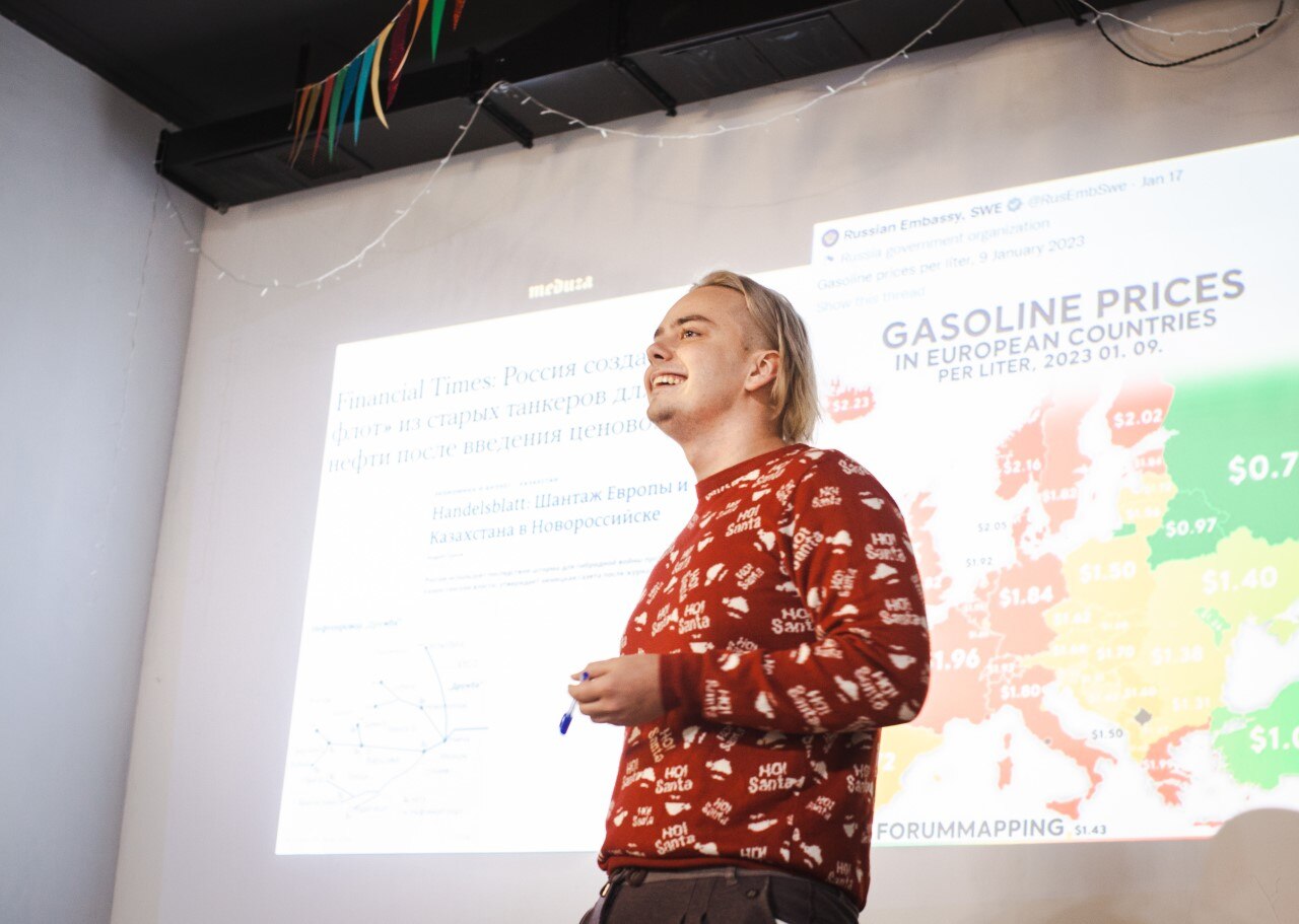 A man in a patterned red jumper stands on a stage with a presentation on gas prices projected behind him
