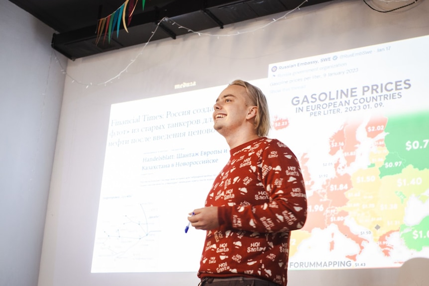A man in a patterned red jumper stands on a stage with a presentation on gas prices projected behind him