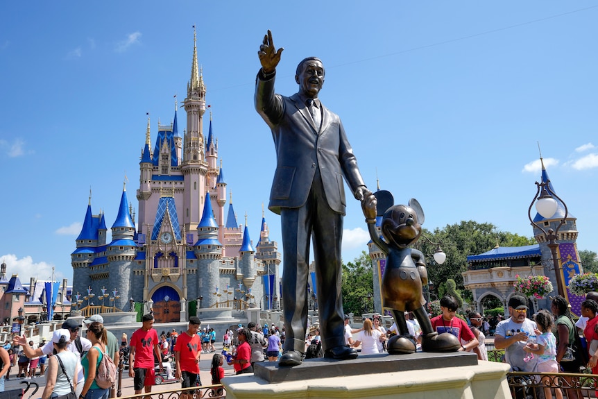 Guests at a Disney park pass a statue of Walt Disney holding the hand of Mickey Mouse.