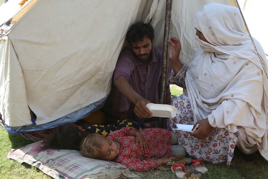 A man, a woman and two children sit in the shade of a small tent