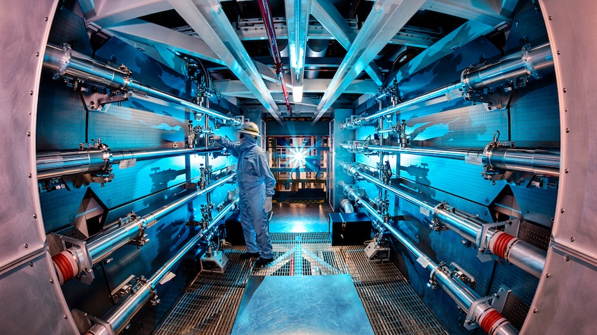 Person in protective suit inside chamber with pipes and blue light.