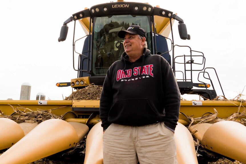 A clean-shaven man stands in front of a yellow corn harvester.