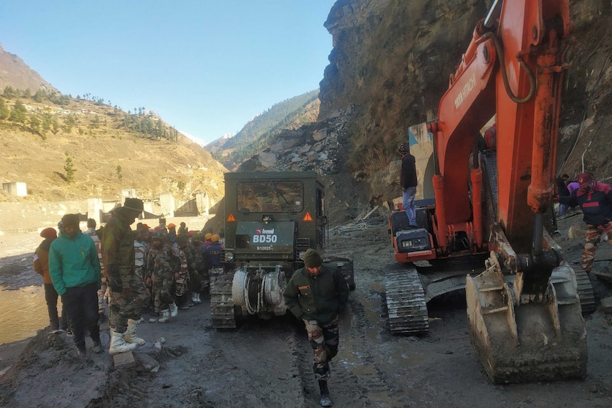 Rescuers move heavy earthmoving equipment along a muddy track in a long valley.