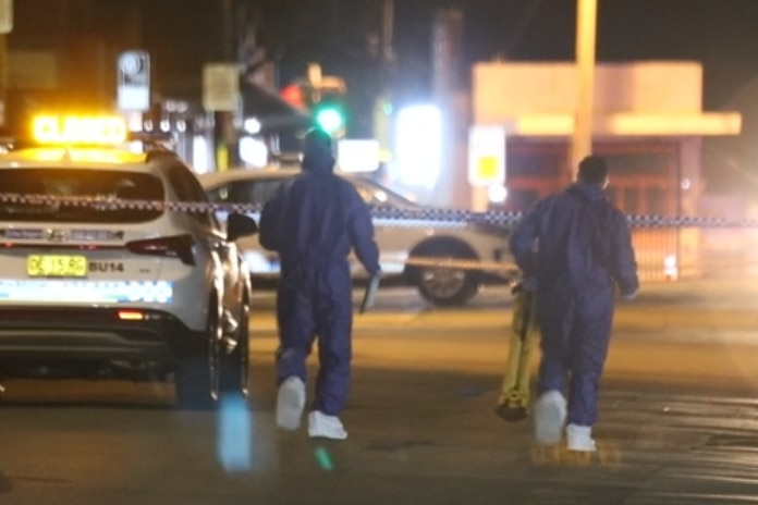 Two people wearing blue jumpsuits walk toward police tape across a road
