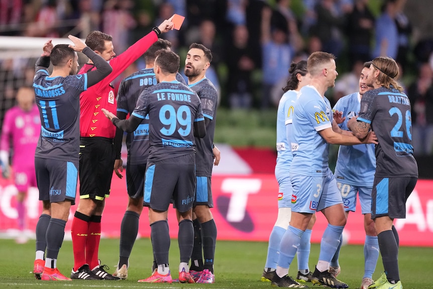 A Sydney FC A-League player hs shown a red card by the referee as players surround the match official.