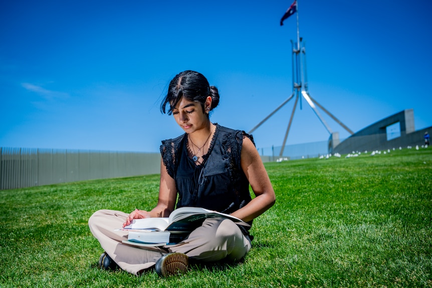 A woman in black top, light trousers, sits on bright green grass reading text book, the Australian flag over parliament behind