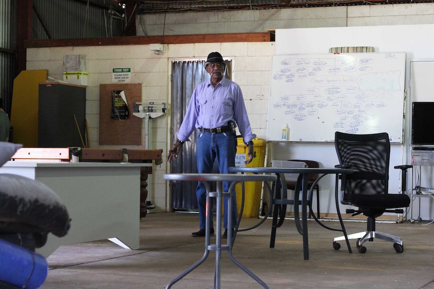 An old man stands in a shed filled with equipment.