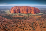 This aerial view of Uluru shows a large red rock in a scrubby desert landscape