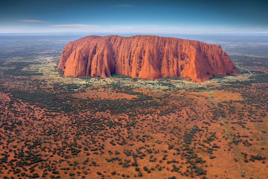 This aerial view of Uluru shows a large red rock in a scrubby desert landscape