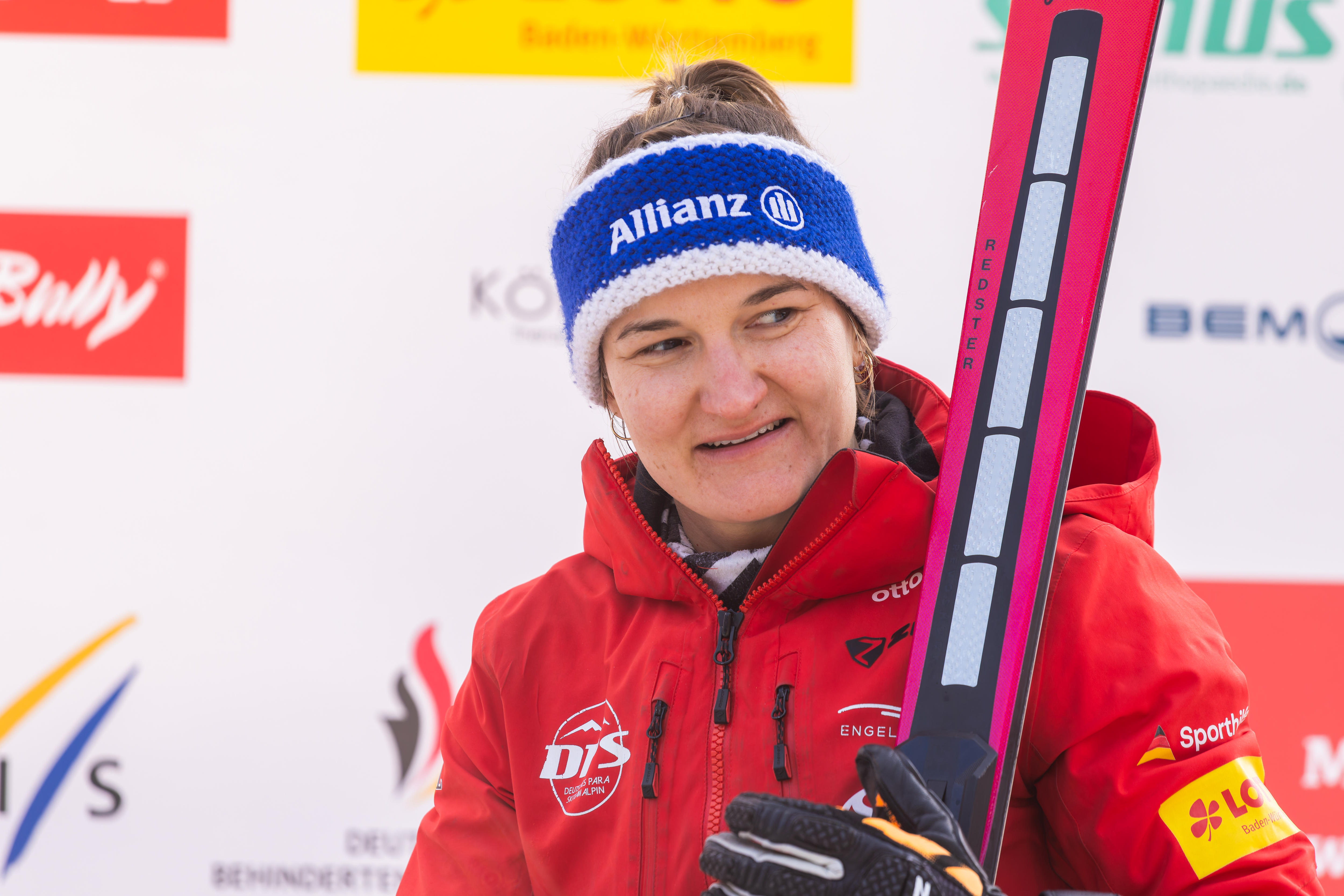 Uma mulher está sorrindo em frente a uma parede branca, segurando alguns esquis e vestindo uma jaqueta vermelha de inverno e uma faixa azul na cabeça