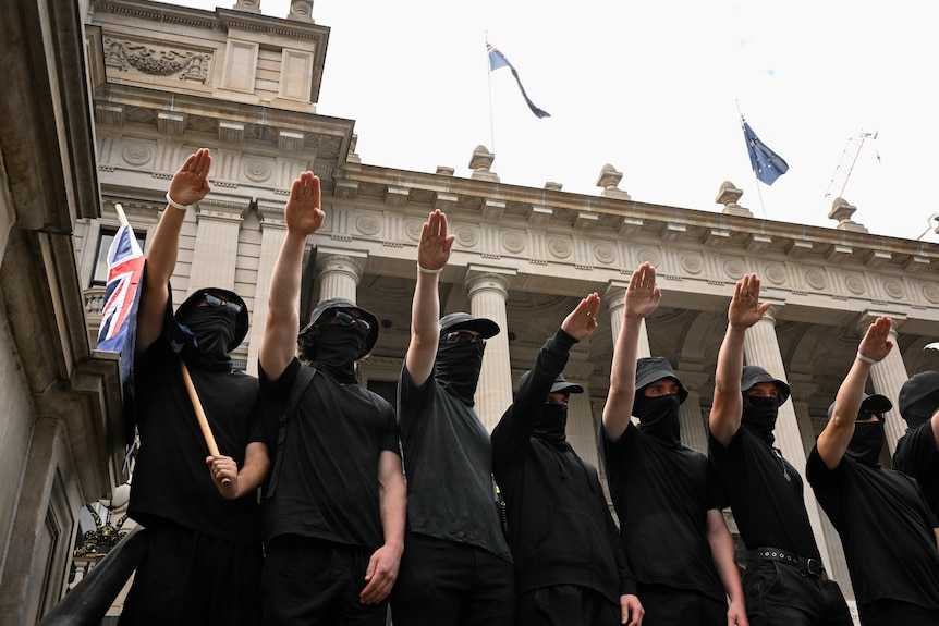Men wearing masks and gats hold their hands up in an apparent Nazi salute, with Victorian Parliament behind them.