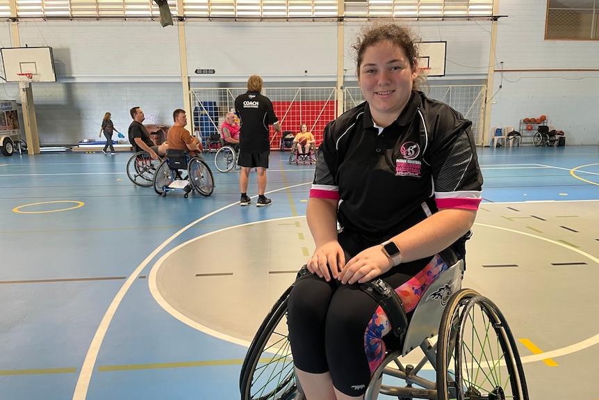 A young women in a wheelchair smiles on a basketball court with players behind her 