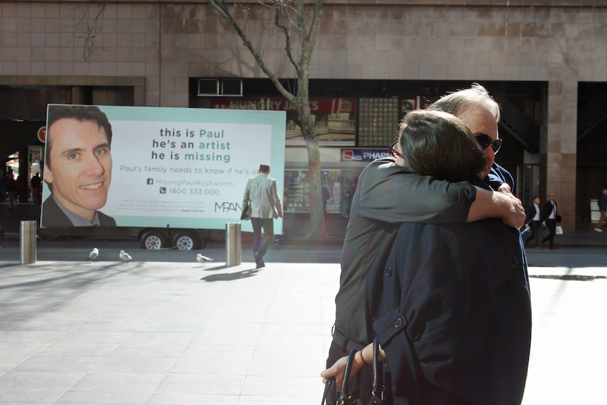 Two people hug in front of a billboard of a missing person. 