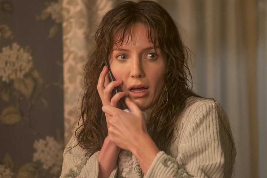 A woman with unruly long wet hair holds a phone up to her ear, her mouth is open in fear