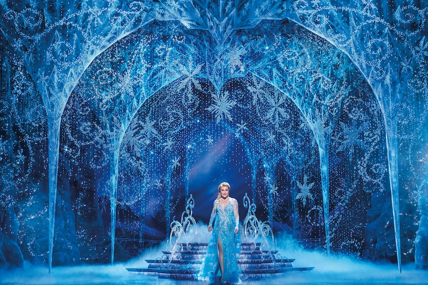On stage a woman in blue dress and tiara stands in an icey palace