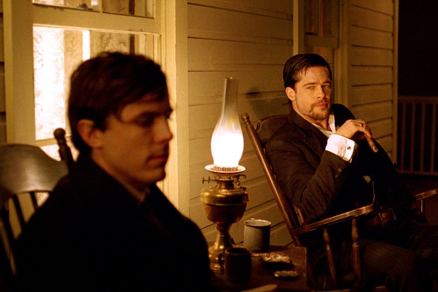 Brad Pitt looks at a blurred Casey Affleck, as they sit together illuminated by yellow light on a front porch, in period costume