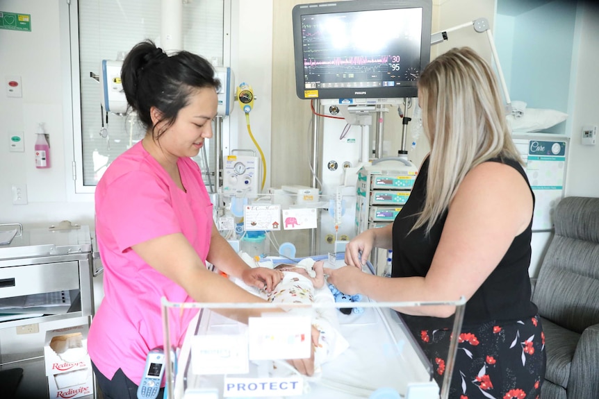 A nurse examines a small baby in a hospital bed as another woman looks on.