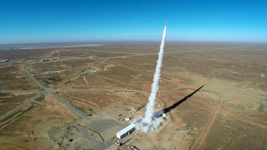 An atmospheric photo shows a rocket below launched with a vertical column of smoke behind it falling toward brown landscape.